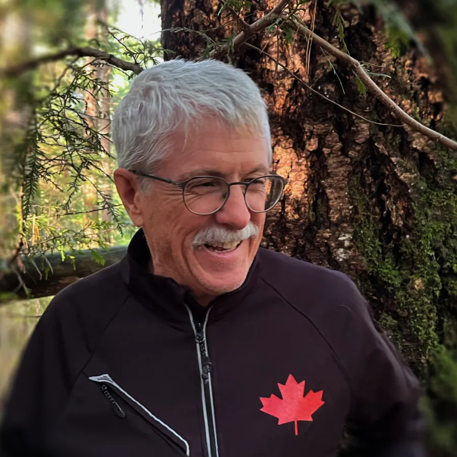 Image of author R. E. St. Denis in a BC forest wearing and black jacket with a red Canadian maple leaf